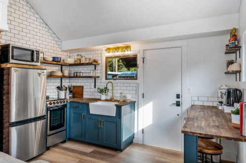 Modern kitchen with blue cabinets, white tiles, stainless steel appliances, and wooden shelves, featuring a cozy dining area.