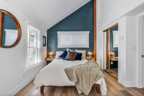 Cozy bedroom with a bed, blue accent wall, round mirror, and natural light from windows. Warm, inviting decor.