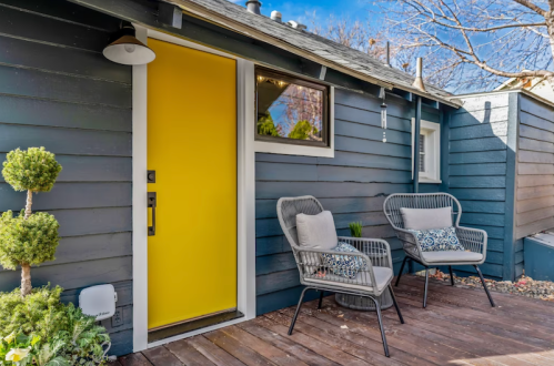 A cozy entrance with a bright yellow door, two chairs, and greenery against a dark blue wooden exterior.
