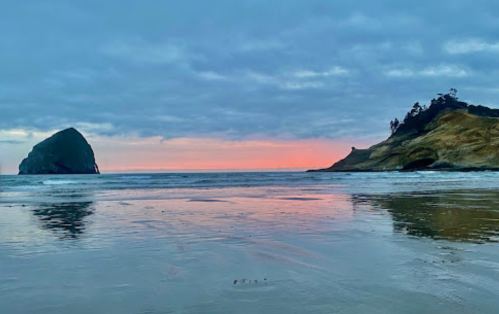 A serene beach at sunset, featuring a large rock formation and gentle waves reflecting the colorful sky.