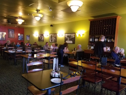A cozy restaurant interior with diners at tables, warm lighting, and wine bottles displayed on a shelf.
