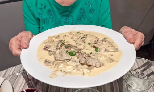 A person holds a large plate of creamy mushroom sauce with meat, garnished with parsley, on a dining table.