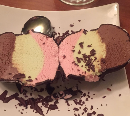 A sliced dessert featuring layers of chocolate, strawberry, and vanilla ice cream, coated in chocolate shavings.
