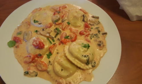 A plate of ravioli in a creamy sauce, topped with cherry tomatoes, mushrooms, and fresh herbs.