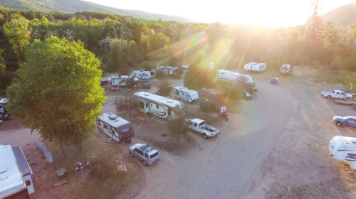 Aerial view of a campground with RVs and trucks surrounded by trees and mountains at sunset.