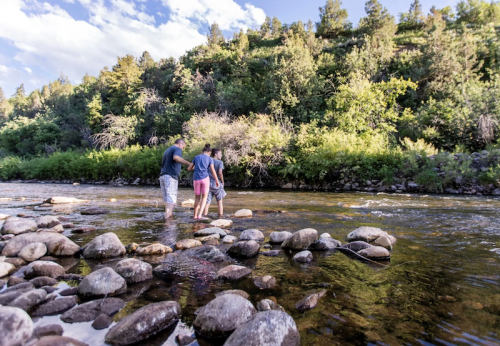 Two people wade through a rocky river, surrounded by lush greenery and a blue sky with scattered clouds.