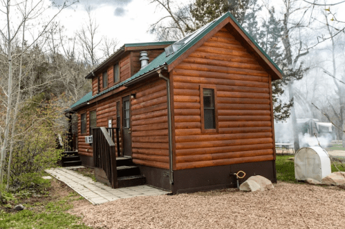 A cozy wooden cabin with a green roof, surrounded by trees and a stone pathway.