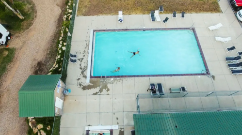 Aerial view of a swimming pool with two people swimming, surrounded by lounge chairs and grassy areas.
