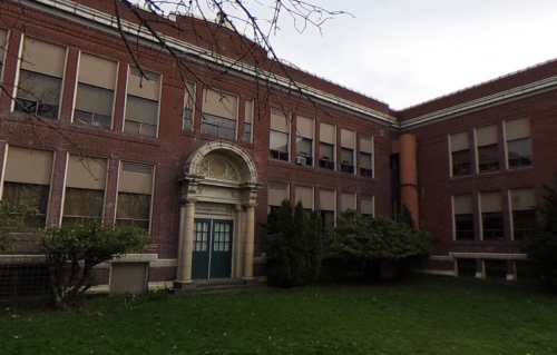 A brick school building with large windows and a green entrance, surrounded by grass and small trees.