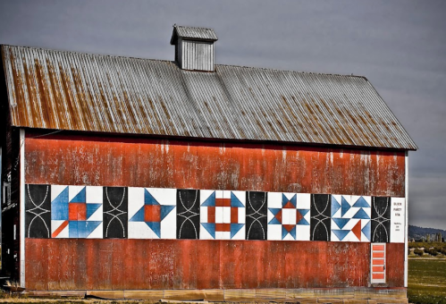 A weathered red barn with a metal roof, featuring colorful geometric quilt patterns painted on its side.