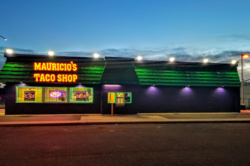 A brightly lit taco shop with a green roof and neon signs, set against a twilight sky.