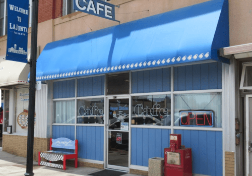 A blue and white café with an "OPEN" sign, featuring a striped awning and a red bench outside.