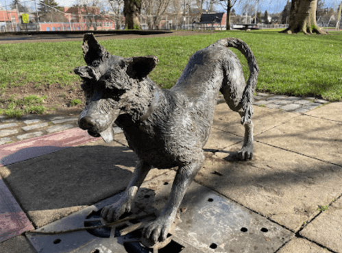 A bronze sculpture of a dog in a playful stance, set on a stone pathway in a grassy park.