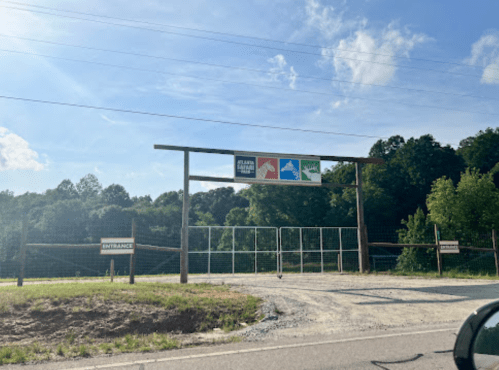 A wooden entrance sign with colorful icons, surrounded by trees and a gravel path, indicating a park or recreational area.