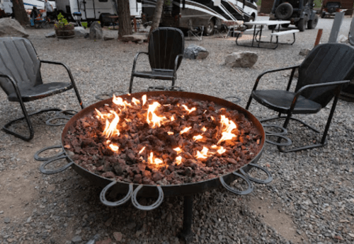A fire pit surrounded by chairs, with flames flickering over rocks in a gravel campsite setting.