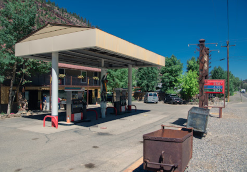 A gas station with vintage pumps, surrounded by trees and buildings, on a sunny day.