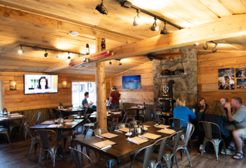 Cozy wooden restaurant interior with tables, a TV, and patrons enjoying their meals. Warm lighting and rustic decor.