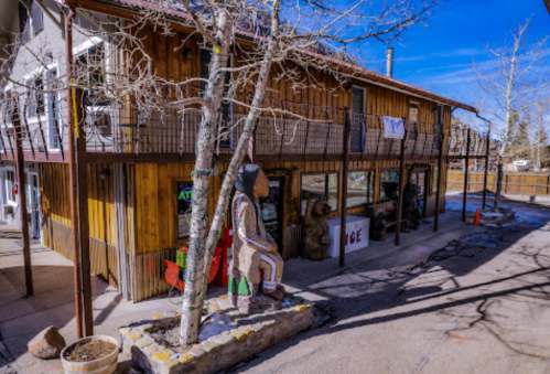 A rustic wooden building with a balcony, featuring sculptures and a tree in front, under a clear blue sky.