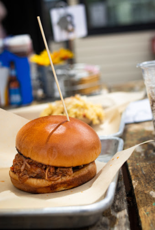 A pulled pork sandwich on a bun with a toothpick, served on a tray with sides in a casual dining setting.