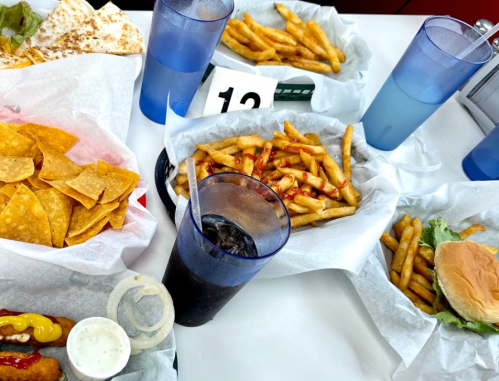 A table with various fast food items: fries, nachos, a burger, and drinks in blue cups, with a number 12 marker.