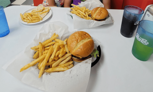 Two plates of burgers with fries on a table, accompanied by colorful drinks in plastic cups.