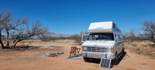 A silver camper van parked in a desert landscape with a solar panel and a chair nearby under a clear blue sky.