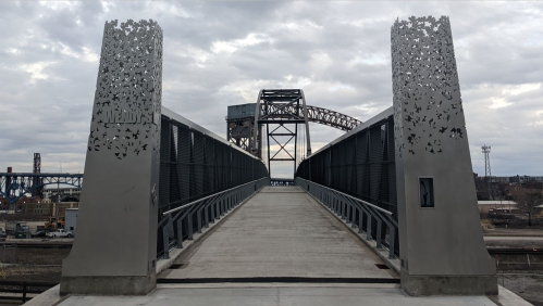 A modern bridge with decorative metal pillars, leading to a raised section and cloudy sky in the background.