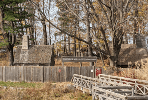 A rustic scene of Pioneer Village with wooden buildings and a sign, surrounded by trees and autumn foliage.