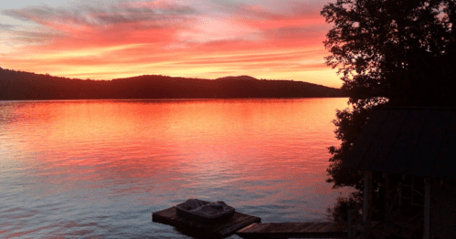 A serene lake at sunset, with vibrant orange and pink hues reflecting on the water and silhouettes of trees in the background.