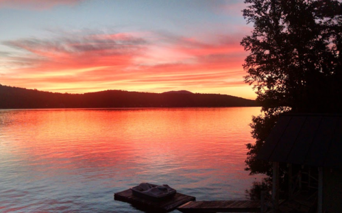 A serene lake at sunset, with vibrant orange and pink skies reflecting on the water, surrounded by trees and mountains.