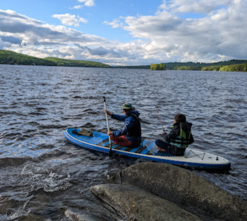 Two people paddle on a stand-up paddleboard in a lake, surrounded by trees and cloudy skies.