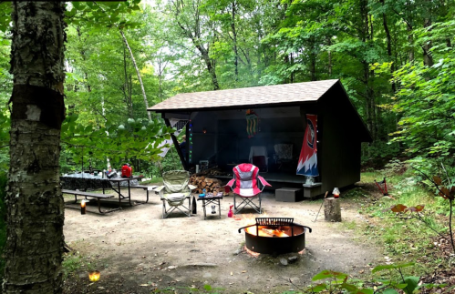 A cozy campsite with a cabin, picnic table, chairs, and a fire pit surrounded by trees.