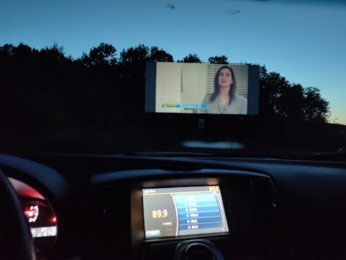 A car interior at dusk, showing a drive-in screen with a woman speaking on a news program. Radio display visible.