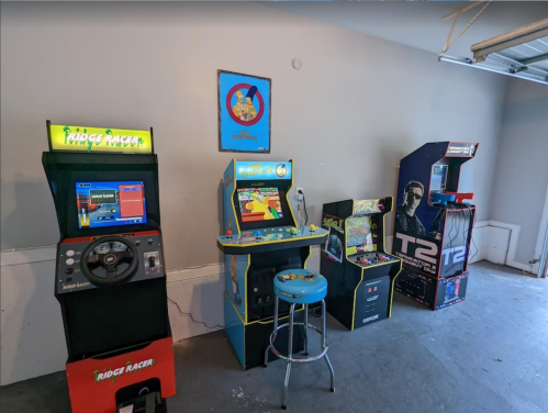 A garage with four arcade machines, including Ridge Racer and T2, alongside a blue stool and a framed poster on the wall.