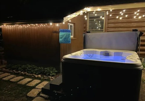 A lit hot tub at night beside a wooden cabin, with string lights and a towel hanging nearby.