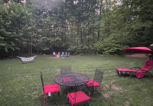 A serene backyard scene with a round table and chairs, a hammock, and lounge chairs surrounded by lush greenery.