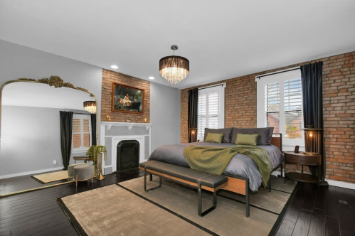 A stylish bedroom featuring a brick wall, large mirror, chandelier, and cozy bedding with green accents.