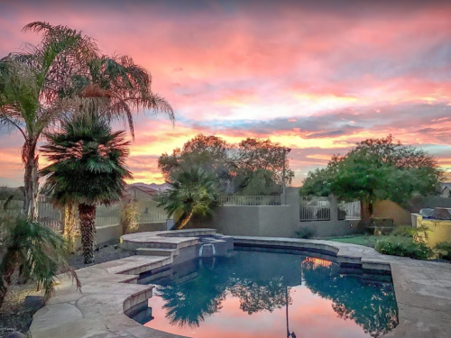 A serene backyard pool at sunset, surrounded by palm trees and vibrant pink and orange skies.