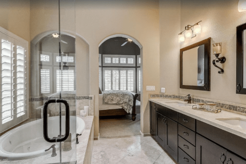 Spacious bathroom with a glass shower, large tub, dual sinks, and a view of a bedroom through arched doorways.