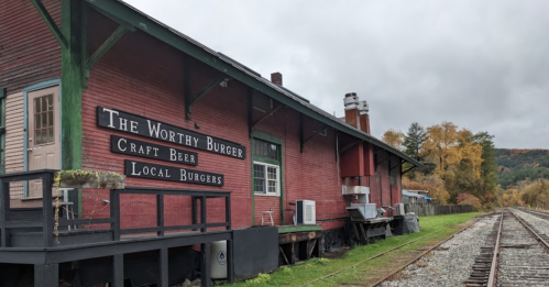 A red building with a sign reading "The Worthy Burger" next to train tracks, surrounded by autumn foliage.