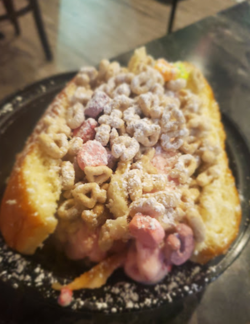 A dessert sandwich filled with ice cream, topped with cereal and powdered sugar, served on a toasted bun.