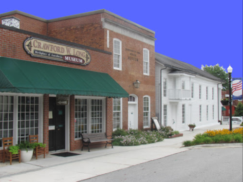Exterior view of the Crawford W. Long Museum, featuring brick and white buildings with a green awning and flower beds.