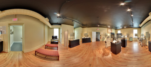A panoramic view of a museum exhibit featuring display cases and seating in a well-lit room with wooden floors.