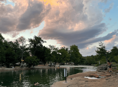 A serene park scene with a pond, trees, and people enjoying the outdoors under a colorful sky with clouds.