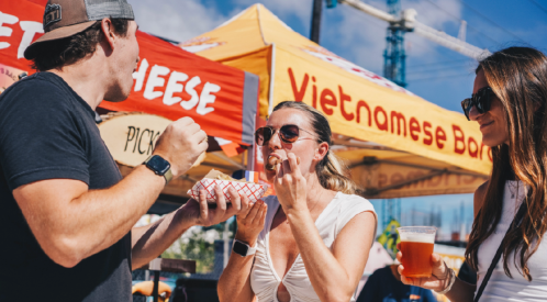 A group of three people enjoying food and drinks at a vibrant outdoor market with colorful vendor tents.