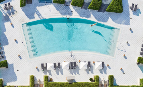 Aerial view of a curved swimming pool surrounded by lounge chairs and greenery. A person is swimming in the pool.