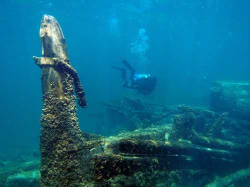 A diver explores a submerged shipwreck, with coral and marine life covering the remains in clear blue water.