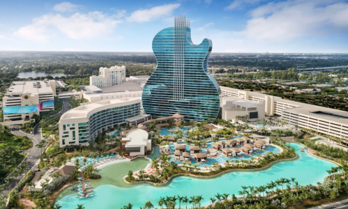 Aerial view of a guitar-shaped hotel with a pool and palm trees, surrounded by a resort and cityscape.