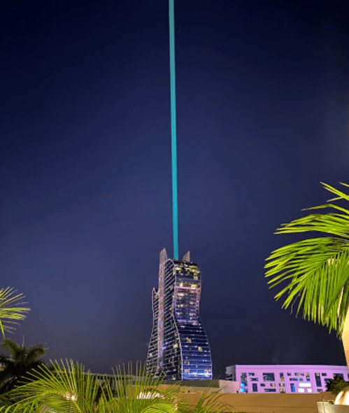 A tall, modern building at night with a bright blue beam of light shooting into the sky, surrounded by palm trees.