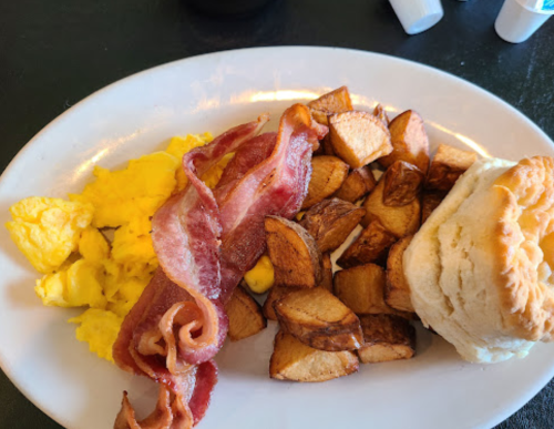 A plate of breakfast featuring scrambled eggs, crispy bacon, seasoned potato wedges, and a biscuit.